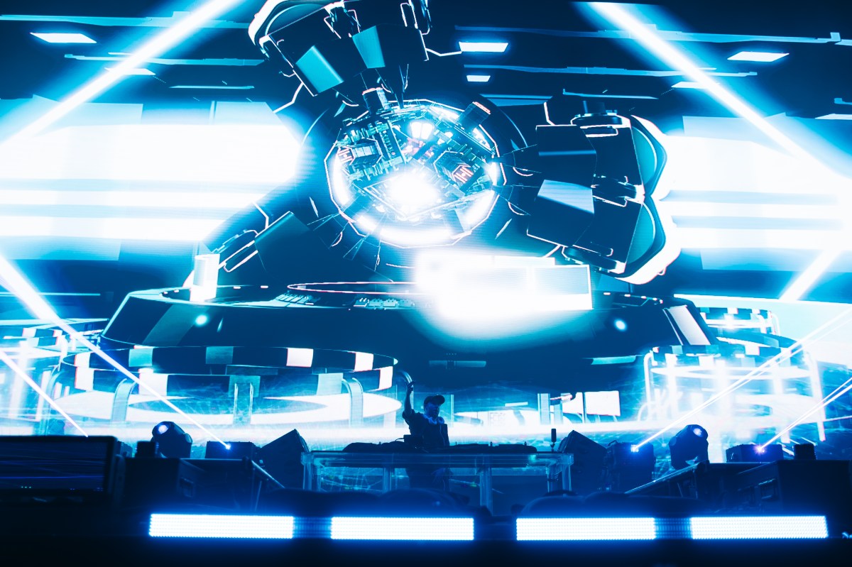 A silhouetted Excision raises a hand in the air in front of a screen with blue techno design.