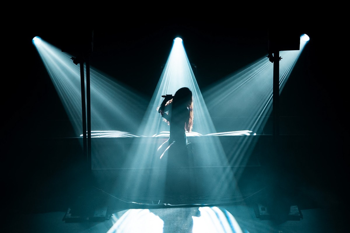 A silhouetted Clara La San perches on a stool, singing into a microphone, while three lights come from directly above her and the left and right.
