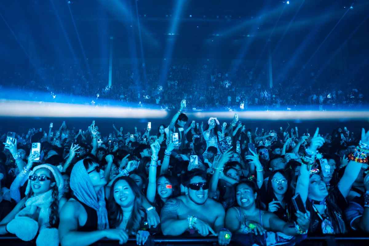 Blue-tinted wide shot of the crowd with their hands up. I beam of horizontal light intersects the middle of the image.